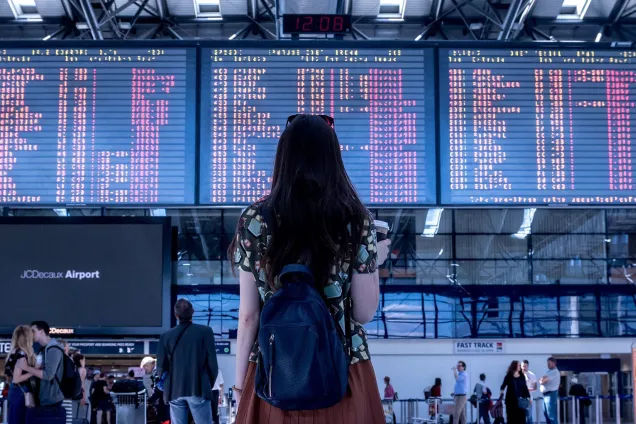 A woman at an airport, looking at the departure flights screen. Photo.