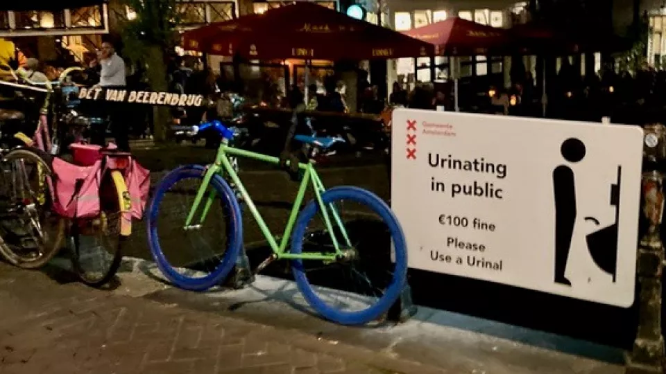 En skylt sitter på ett staket som informerar om att man får 100 euro i böter om man urinerar offentligt. Två cyklar är lutade mot staketet. Foto.