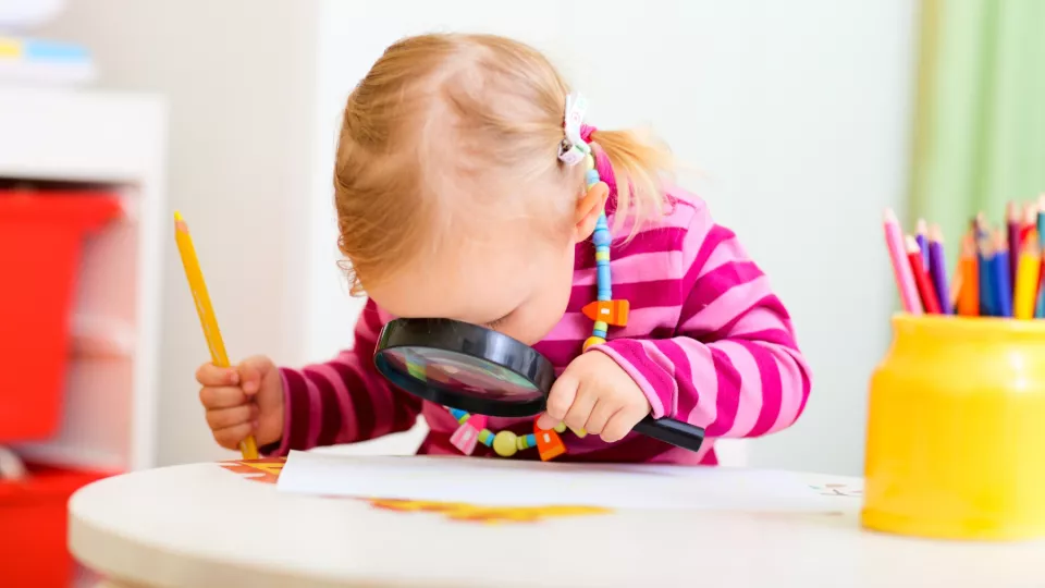 Ett barn sitter vid ett bord med ett förstoringsglas i ena handen och en penna i andra handen. Foto.