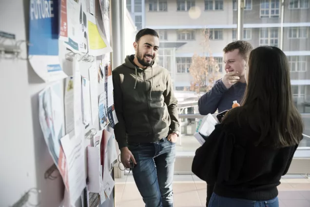 Tre studenter i samtal vid en anslagstavla. En student lutar sig mot tavlan. Foto.