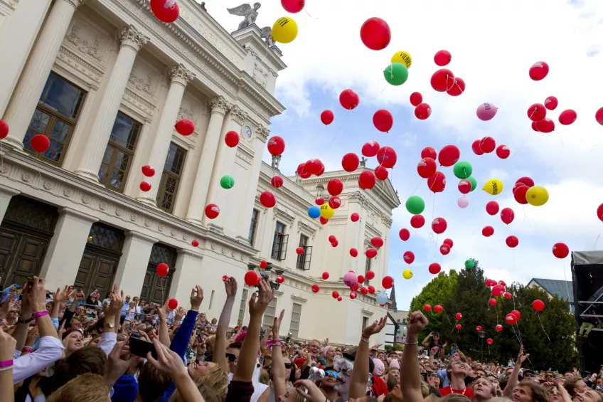 En massa färgglada ballonger släpps upp i luften av en folksamling framför universitetshuset. Foto.